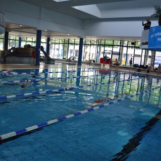 Schwimmbecken mit mehreren Bahnen, Schwimmern und einer Zuschauertribüne, im Hintergrund eine Rutsche.