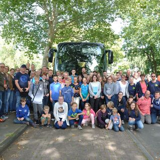Gruppenfoto von Kindern und Erwachsenen vor einem Bus, umgeben von Bäumen auf einer Straße.