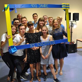 Gruppe von Menschen feiert 100 Jahre SCS Schwimmen mit einem bunten Fotorahmen in einer Veranstaltungshalle.