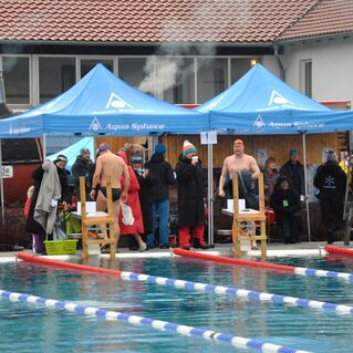 Zwei Schwimmer stehen an Startblöcken an einem Schwimmbecken, während Zuschauer und Teilnehmer am Beckenrand warten.