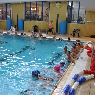 Kinder in Badekleidung stehen am Beckenrand eines Schwimmbads, während andere schwimmen; einige tragen Schwimmkappen.