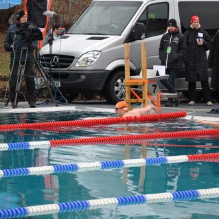Zwei Schwimmer in Badebekleidung schwimmen im Wettkampfbecken, während Zuschauer und Kameraleute zuschauen.