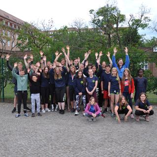 Gruppenerfoto von Jugendlichen und Erwachsenen im Freien, alle mit erhobenen Händen vor einem Gebäude und Bäumen.