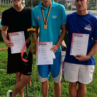 Drei Personen stehen lächelnd auf einer Holzplattform, halten Urkunden und einen Sonnenblumenstrauß in der Mitte.