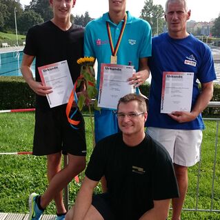Vier Personen stehen und sitzen zusammen, halten Urkunden und eine Sonnenblume in einer Außenumgebung.