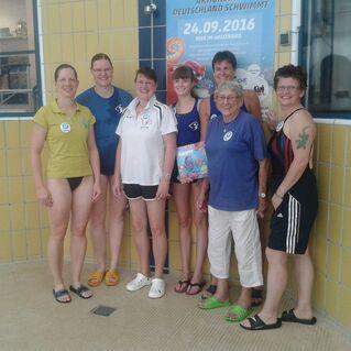 Gruppe von sieben Frauen in Badebekleidung vor einer Wand mit einem Plakat im Schwimmbad, Lächeln und freundliche Stimmung.