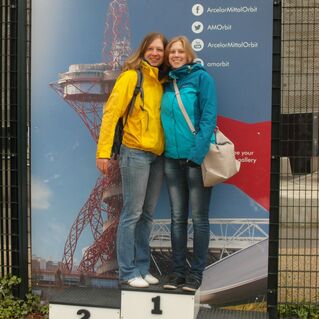 Zwei Frauen posieren auf Podesten mit den Nummern 1, 2 und 3 vor einer Wand mit einem Bild des ArcelorMittal Orbit.