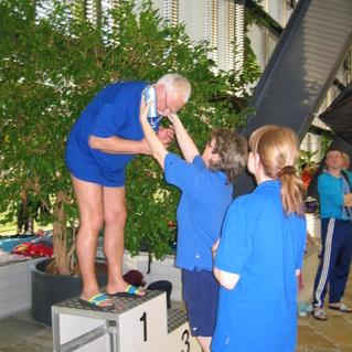 Ein älterer Mann steht auf einem Siegertreppchen, während drei Personen in blauen Shirts ihm applaudieren.