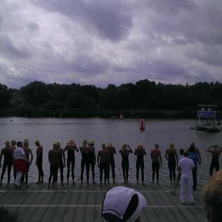 Gruppe von Schwimmern in Neoprenanzügen mit Badekappen am Ufer eines Gewässers vor einem Wettkampf.