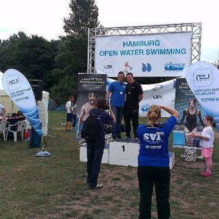 Zwei Schwimmer stehen auf dem Wettkampfsiegertreppchen bei den Hamburger Open Water Swimming, umgeben von Zuschauern.