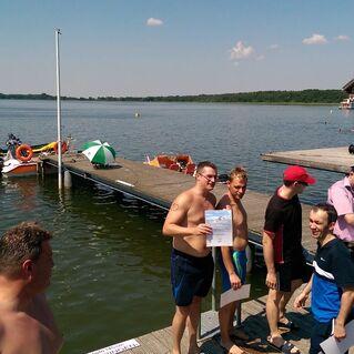 Gruppierung von Menschen am Ufer eines Sees, einige halten Urkunden, Boote und eine Holzplattform im Hintergrund.