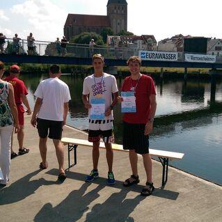 Zwei Männer halten Urkunden am Ufer eines Gewässers, im Hintergrund eine Menschenmenge auf einer Brücke.