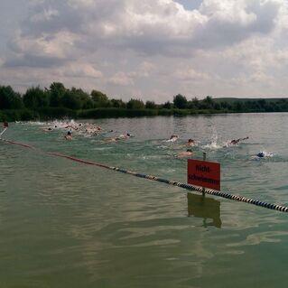 Schwimmer in einem Badesee, mit „Nichtschwimmer“-Schild nahe einer Absperrung und Menschen am Ufer.