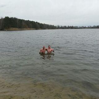 Zwei Männer stehen nackt im Wasser eines Sees, umgeben von Bäumen und bewölktem Himmel.