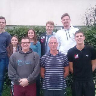 Gruppenfoto von acht Personen vor einer grünen Hecke, einige lächeln, andere schauen frontal.