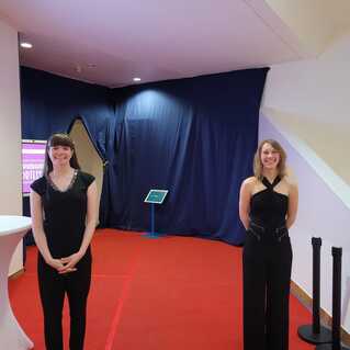 Zwei Frauen in eleganter Kleidung stehen vor einem rot Teppich in einem Eingangsbereich mit blauer Vorhang im Hintergrund.