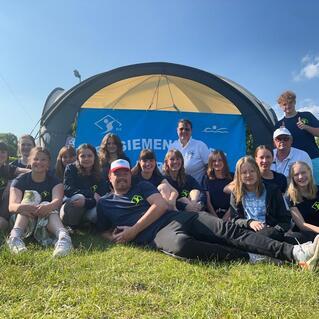 Gruppe junger Sportler und Trainer posiert lächelnd vor einem Zelt mit dem Banner "Siemens" auf einer Wiese.