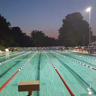 Beleuchteter Schwimmbecken mit mehreren Bahnen und Zuschauern bei Dämmerung. Ein Schwimmer ist im Wasser sichtbar.