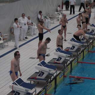 Schwimmer in Badebekleidung stehen an Startblöcken neben einem Schwimmbecken bei einem Wettkampf.