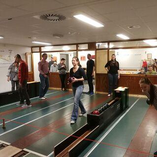 Gruppe von Personen in einer Bowlingbahn, einige halten Bowlingkugeln, andere beobachten das Spiel.