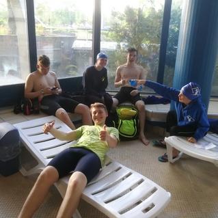 Vier Jugendliche im Schwimmbad sitzen entspannt auf Liegen, während einer von ihnen eine Wasserflasche hält und lächelt.