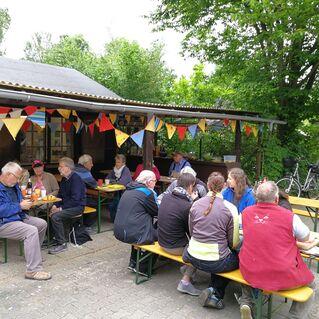 Gruppe von Menschen sitzt an Bänken unter einem Vordach, umgeben von bunten Fähnchen und Bäumen.