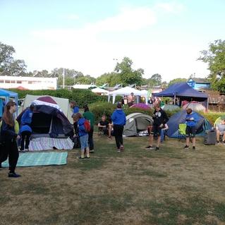 Campingplatz mit mehreren Zelten und Personen, die sich bewegen und miteinander kommunizieren, unter blauem Himmel.