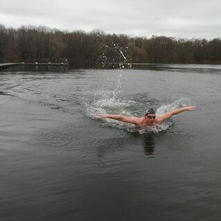 Ein schwimmender Mann in einer grauen Badekappe und Sonnenbrille macht einen Schwimmzug im kalten, stillen Wasser.