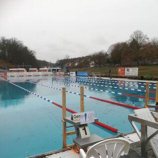 Schwimmbecken mit Startblöcken und bunten Bahnmarkierungen, umgeben von einer leeren Tribüne und einem grauen Himmel.