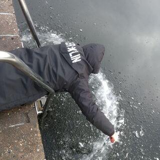 Person in schwarzer Jacke beugt sich über eine gefrorene Wasseroberfläche und bricht das Eis mit der Hand.