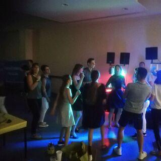 Gruppe von tanzenden Personen in einem beleuchteten Raum mit verschiedenen bunten Lichtern und Lautsprechern.