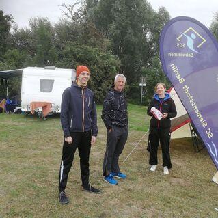 Drei Personen stehen auf einem Campingplatz neben einem Wohnwagen und einem Zelt, während ein Fahne im Wind weht.