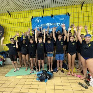Gruppe von Jugendlichen in Schwimmbekleidung hebt begeistert die Hände vor einem Banner des SC Siemensstadt Berlin.