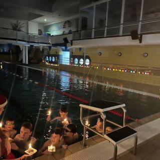 Gruppe von Kindern im Wasser mit Kerzen, während ein Erwachsener mit Weihnachtsmütze ihnen zuschaut. Beleuchtetes Schwimmbad im Hintergrund.