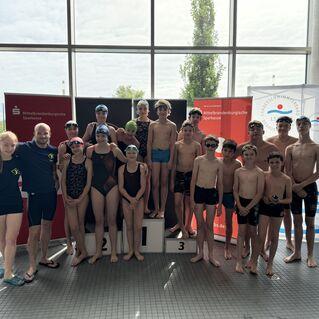 Gruppe von Schwimmern in Badeanzügen, die auf einem Siegertreppchen stehen, mit Sponsorenbanner im Hintergrund.