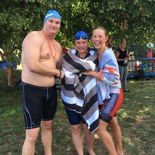 Drei Schwimmer stehen lächelnd im Freien, zwei mit Badekappen und Handtüchern umwickelt, umgeben von Bäumen.