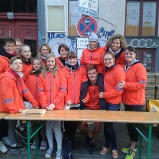 Gruppe von 14 Personen in orangenen Jacken vor einem Gebäude, Stehtisch mit Holzplatten und Bananenkisten im Hintergrund.