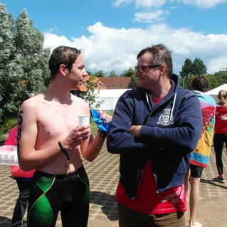 Zwei Personen sprechen am Rande einer Veranstaltung, während Schwimmer im Hintergrund vorbeigehen. Es ist sonnig.