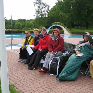 Fünf Personen sitzen auf Campingstühlen am Rand eines Schwimmbeckens in einem Park. Grüne Bäume im Hintergrund.