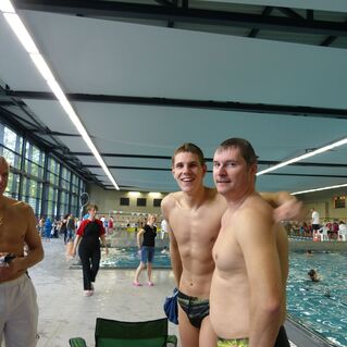 Drei Männer stehen am Rand eines Schwimmbeckens, lächeln und sind in Badekleidung gekleidet. Im Hintergrund sind Schwimmer zu sehen.
