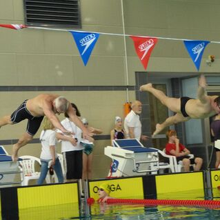 Zwei Schwimmer springen gleichzeitig von Startblöcken ins Wasser, während Zuschauer am Beckenrand zuschauen.