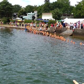 Gruppenschwimmen mit Kindern in Orangekappen an der Uferkante, Zuschauer stehen auf dem Steg und am Rasenbereich.