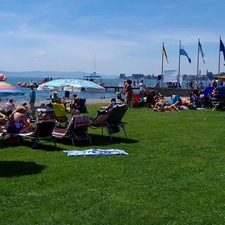Blick auf einen Strand mit Sonnenliegen, Sonnenschirmen und Menschen, die sich am Wasser und auf der Wiese aufhalten.