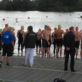 Schwimmer in Neoprenanzügen stehen am Ufer eines Gewässers, bereit für einen Wettkampf. Im Hintergrund schwimmen Boote.