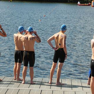 Gruppe von Schwimmern in Badekappen mit nummerierten Rücken steht am Ufer eines Gewässers.