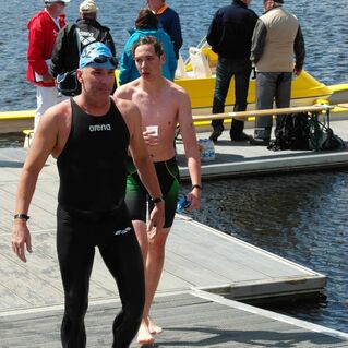 Zwei Schwimmer in Neoprenanzügen gehen auf einen Steg am Wasser, während Zuschauer im Hintergrund stehen.