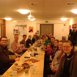 Gruppenevent mit mehreren Personen an einem langen Tisch, gedeckt mit verschiedenen Speisen und Getränken in einem Raum.