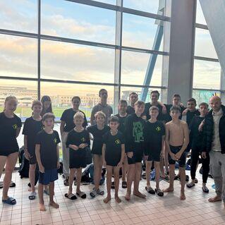 Gruppenshot von Schwimmern in schwarzen Shirts, stehend vor großen Fenstern in einem Sportzentrum.