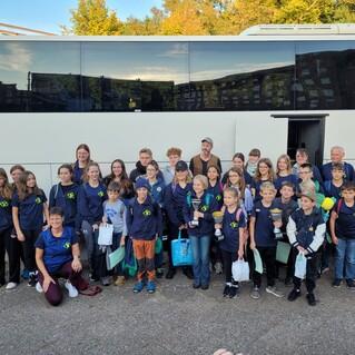 Gruppe von Schülerinnen und Schülern mit Pokalen vor einem Reisebus in einer wolkenlosen Umgebung.