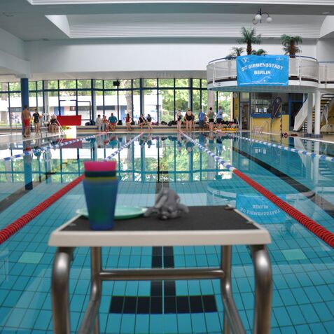 Blick auf ein Schwimmbad mit einer Tischtennisplatte und bunt gestapelten Bechern im Vordergrund. Schwimmer warten am Beckenrand.